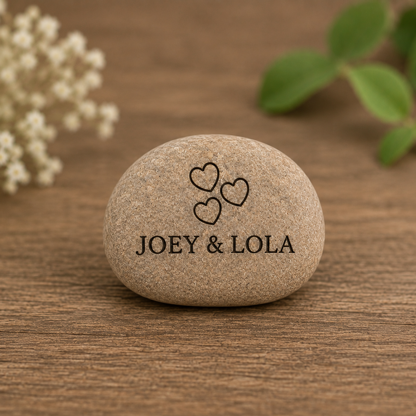 Stone with 'JOEY & LOLA' and heart symbols on a wooden surface with flowers and leaves.
