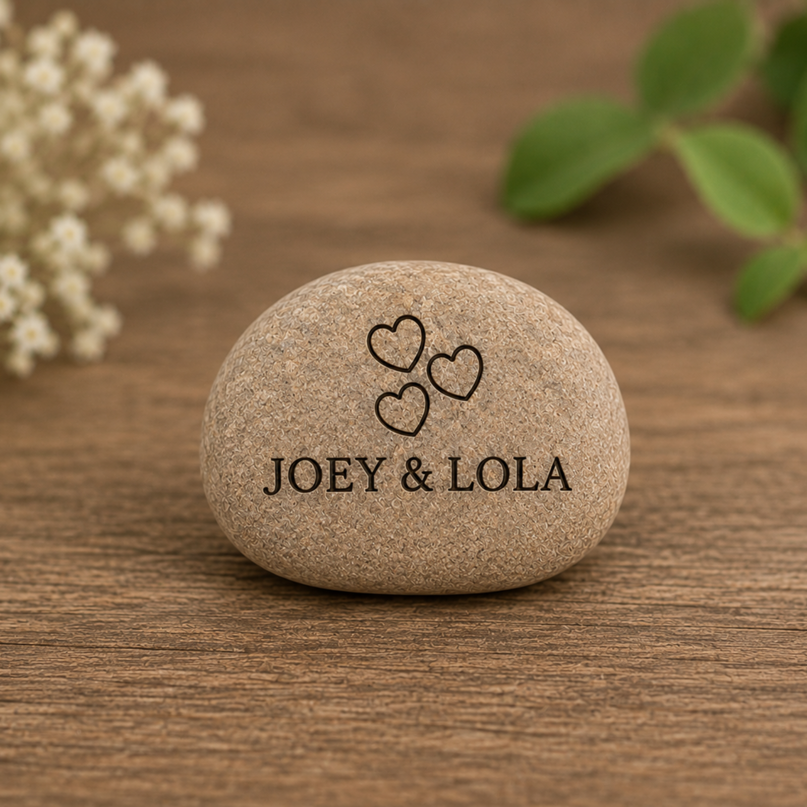 Stone with 'JOEY & LOLA' and heart symbols on a wooden surface with flowers and leaves.